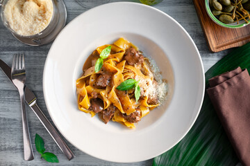 Papardelle pasta with beef, gravy, parmesan and basil on a white plate. Italian dish served on a table, top view