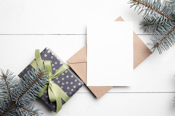 Christmas greeting card mockup with craft paper envelope, green color gift box and fresh fir tree branch on white wooden background, top view, flat lay. Winter holiday card mockup