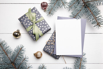 Christmas greeting card mockup with grey envelope, gift boxes and decorations on white wooden background, top view, flat lay. Winter holiday card mockup with fir tree branches and balls