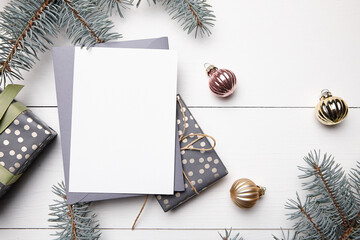 Fototapeta premium Christmas greeting card mockup with grey envelope, gift boxes and decorations on white wooden background, top view, flat lay. Winter holiday card mockup with fir tree branches and balls