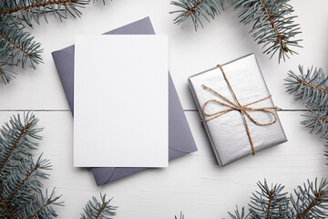 Christmas greeting card mockup with grey envelope, silver color gift box and green fir tree branch on white wooden background, top view, flat lay. Empty winter holiday card