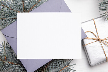 Christmas greeting card mockup with grey envelope, silver color gift box and green fir tree branch on white wooden background, top view, flat lay. Empty winter holiday card