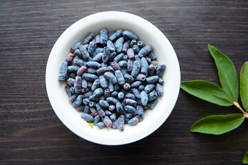 Fresh honeyberry (Lonicera caerulea) in white bowl on wooden table, top view, flat lay. Blue haskap berry with green leaves
