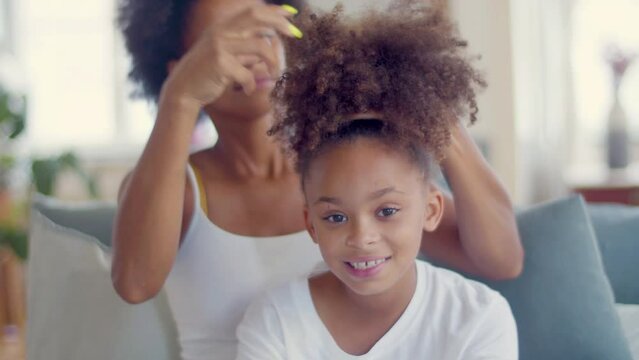 Mother Doing Girls Hair While Cute Black Child Looking At Camera And Smiling. Woman In Background Making Ponytail For Her Daughter. Camera Point Of View. Handheld Shot. Childhood, Happy Family Concept