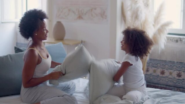 Happy mum and daughter having pillow fight sitting in bed after waking up in the morning. Black woman and little girl having fun, playing and laughing together. Slow motion. Happy family concept.