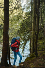 Full length vertical image of tourist woman with big backpack hiking by the river at the forest, enjoying nature view