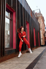 a joyful girl in a red jumpsuit with red hair against the background of urban architecture