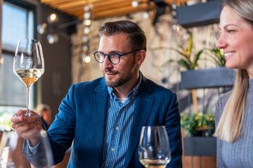 young man sitting at the restaurant with his wife, admiring rich color of his white wine in his glass