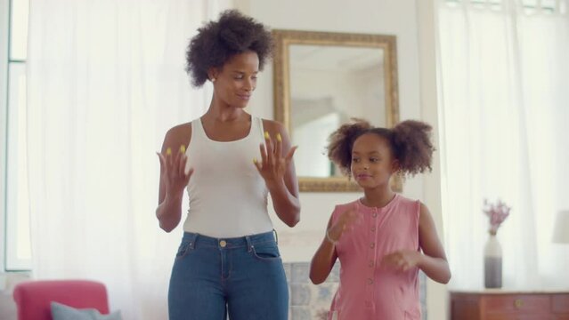 Happy Black mum and daughter dancing at home together, doing morning exercise to music. Smiling cute girl repeating movements after mother. Slow motion. Front view. Leisure, happy family concept.
