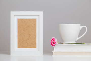 Fototapeta na wymiar White wooden picture frame mockup on desk with books, porcelain mug and beautiful pink flower - composition 