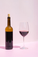 A bottle of red wine and a filled glass next to it in the sun on a pink background. Place for text