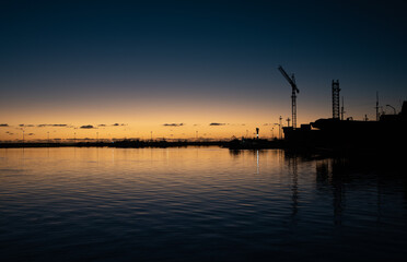 The sun sets on the horizon over the sea. The shadows of an industrial port with cranes can be seen in the foreground. The sea is calm and dark.