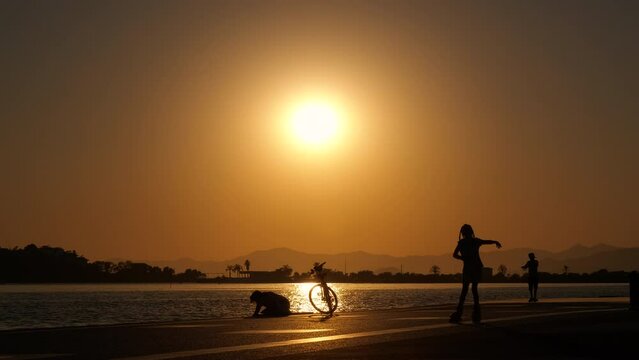 Peoples silhouette by sea at night. Somebody ride a bike, roller skating and strolling silhouette on the city street by the water during sunset.