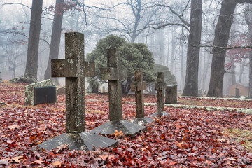 Early foggy morning in a old graveyard with trees and moss covered headstones and four crosses. Autumn leaves covering the ground in Sewanee Tennessee university cemetery.