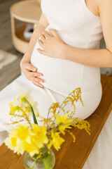 A pregnant woman holding her hands on her stomach, sitting at home on the couch. Close-up.
