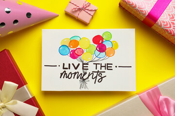 Life-affirming phrase Live The Moments. Flat lay composition with card and gift boxes on yellow background