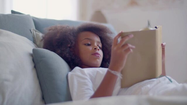 Cute Black girl reading in bed, holding hardcover book in hands. Beautiful child enjoying good story, reading aloud, relaxing in afternoon at home. Dolly medium shot. Childhood, leisure time concept.