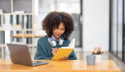 Cute african american female student wearing headphones with afro dreadlocks, studying remotely from home, using a laptop, taking notes on notepad during online lesson, e-learning concept,  - obrazy, fototapety, plakaty