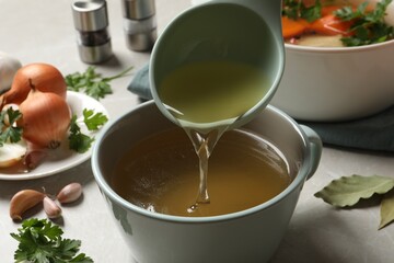Pouring hot delicious bouillon into cup on light grey table, closeup