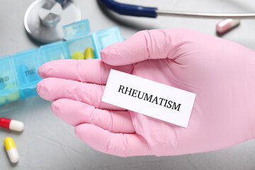 Doctor in glove holding sheet of paper with word Rheumatism above grey table, closeup