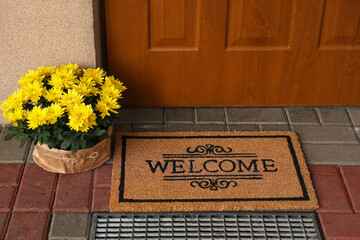 Door mat with word Welcome and beautiful flowers near entrance - obrazy, fototapety, plakaty