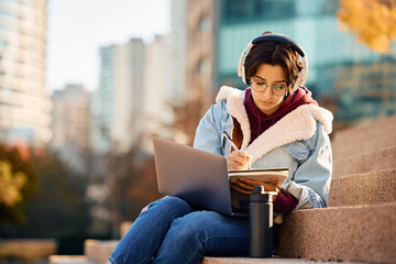 Female student taking notes while e-learning on laptop outdoors at campus.