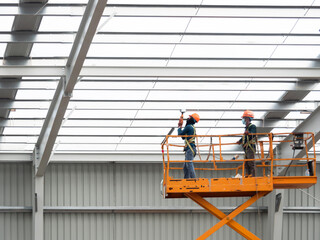 Workers install lighting fixture in a hug industrial warehouse using hydraulic scissor lift. MEP work in construction site.