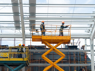 Workers install lighting fixture in a hug industrial warehouse using hydraulic scissor lift. MEP work in construction site.