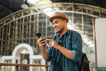 Traveler young asian man using cellphone booking trip at terminal train station. Happy tourist travel by train using smartphone searching location. Male Backpacker arrival at platform railway.