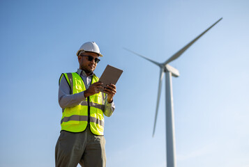 Engineer India man working with tablet at windmill farm Generating electricity clean energy. Wind turbine farm generator by alternative green energy. Asian engineer checking control electric power