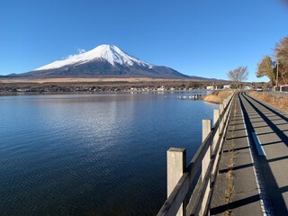 Mount Fuji in the backdrop with a road leading to it and with a clear blue lake on the side.