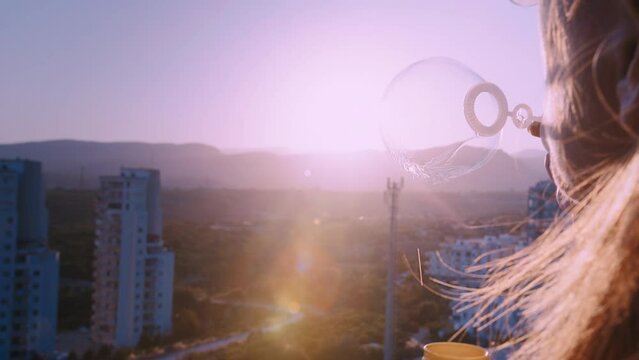 brunette woman blows soap bubbles from a balcony at sunset against the backdrop of colorful mountains, pretty woman inflates soap bubbles and smiles