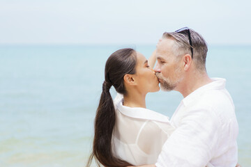 Happy Asian and caucasian white couple enjoy relaxing together at the beach during vacation or their honeymoon dating. Husband gently kissing his wife.