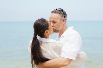 Happy Asian and caucasian white couple enjoy relaxing together at the beach during vacation or their honeymoon dating. Husband gently kissing his wife.