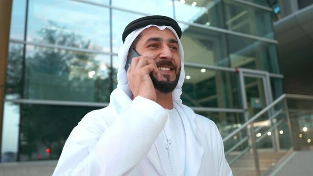 Bearded Man in Kandora talking on the phone at a business area. Arab businessman wearing Dish Dash and Ghutra speaking on smartphone. Communication concept of Middle East people