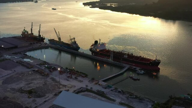 Birds eye view fly around Lumut port industrial park along sungai manjung river with commercial bulk carrier ships docked at the port, tilt up reveals mountain landscape at sunset, Perak, Malaysia.