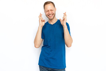 Excited man with crossed fingers standing on white background. Happy Caucasian guy praying for success, squeezing eyes, smiling, hoping for luck. Hope, wish, superstition concept