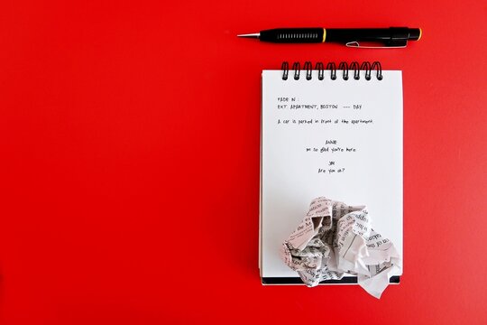 Pen , crumpled paper and  notebook with screenwriting scene written, on red copy space background. Concept of  professional screenwriter job, TV or movie film script writing or writing block   