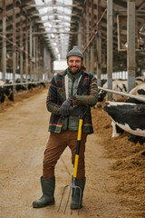 Portrait of young agronomist standing with pitchfork and smiling at camera, he cleaning cowshed in big barn
