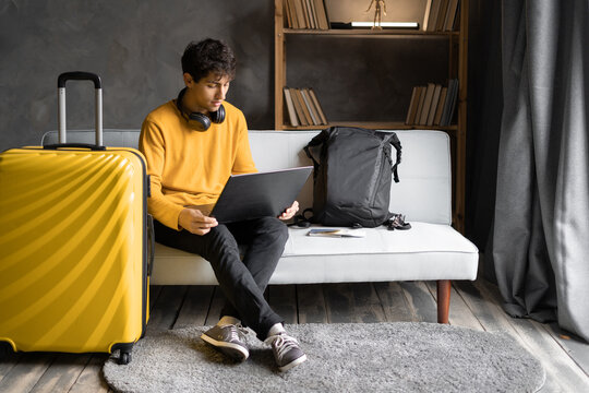 Young man traveler planning vacation trip and using computer to book hotel room online. Male tourist wearing sweater sitting on sofa and browsing rental holiday accommodation website