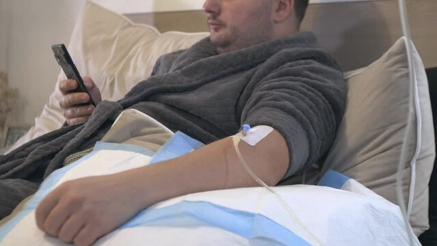 zoom in patient sitting on hospital bed room while receiving a dextrose saline solution, caucasian male chatting on his smartphone with insurance and family member after hospitalisation