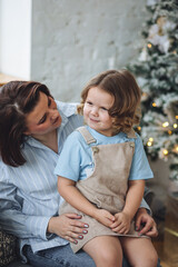 Beautiful mother and her cute little daughter sitting together near Christmas tree, smiling, laughing, hugging. Concept of family love and happiness, motherhood, happy childhood
