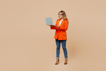 Full body confident young successful employee IT business woman corporate lawyer wear classic formal orange suit glasses work in office hold use laptop pc computer isolated on plain beige background.