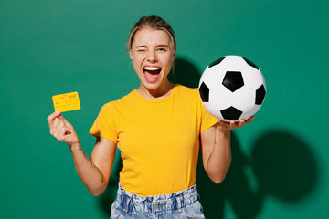 Young woman fan in basic yellow t-shirt cheer up support football sport team hold soccer ball yellow card propose player retire from field watch tv live stream wink isolated on dark green background.