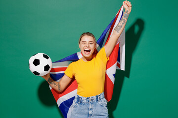 Young fun woman fan wear yellow t-shirt cheer up support football sport team hold in hand big British flag soccer ball watch tv live stream do winner gesture isolated on dark green background studio.