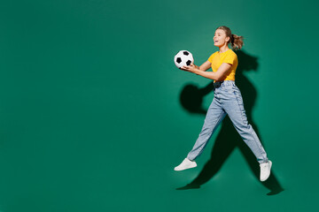 Naklejka na ściany i meble Side view full body young woman fan wear basic yellow t-shirt cheer up support football sport team hold in hand soccer ball watch tv live stream jump high look aside isolated on dark green background.