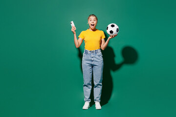 Fototapeta na wymiar Full body young excited woman fan wears basic yellow t-shirt cheer up support football sport team hold soccer ball watch tv live stream use mobile cell phone isolated on dark green background studio.