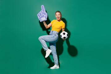 Fototapeta na wymiar Full body side view fun young woman fan wears yellow t-shirt foam 1 glove finger up cheer up support football sport team hold soccer ball watch tv live stream isolated on dark green background studio.