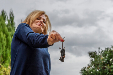 Middle aged blond woman holds a wet rat by the tail with a terrified and disgusted expression