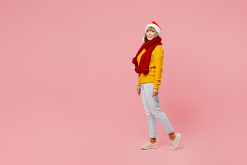 Fototapeta premium Full body side profile view fun elderly woman 50s years old wear yellow knitted sweater red scarf Santa hat posing look camera isolated on plain pink background. Happy New Year Christmas 2023 concept.